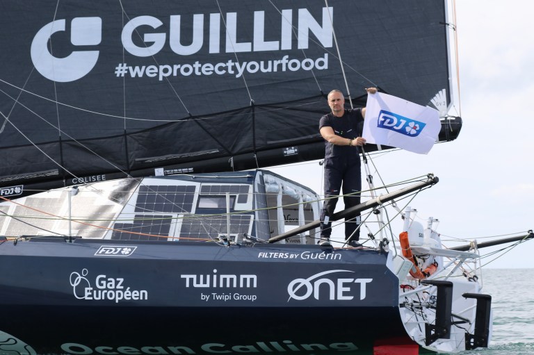 Le skipper Fabrice Amédéo, tenant un drapeau FDJ, sur son IMOCA floqué FDJ